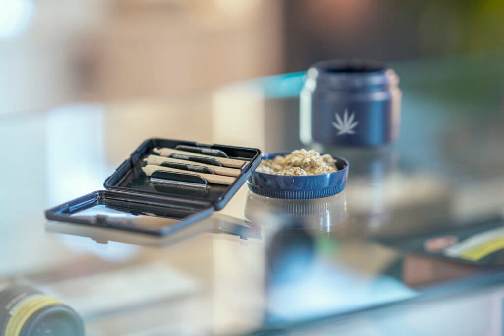 An assortment of cannabis product is laid out and displayed neatly on the counter of a legal retailer.  Joints and cannabis bulbs can be seen laying in their packaging on the glass counter.