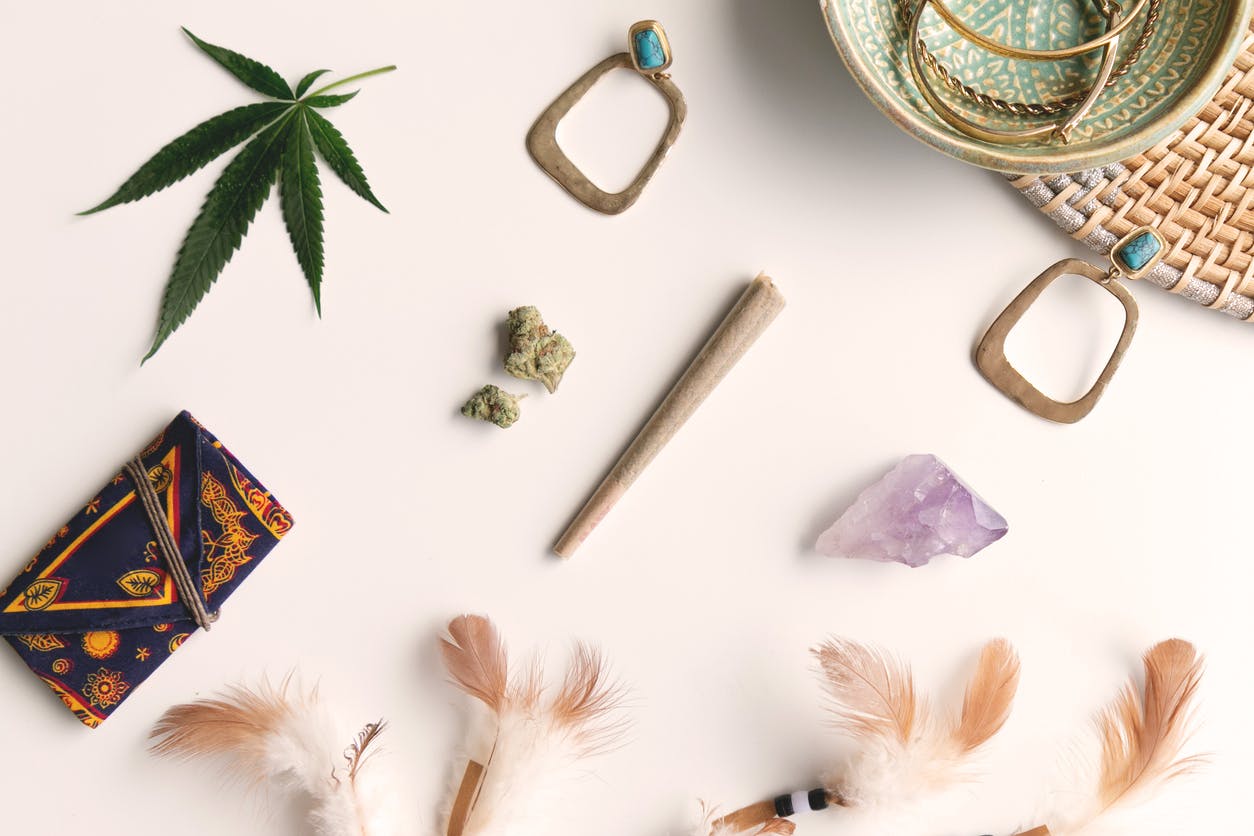 Festival accessories laid out on white and wicker with a joint, nugs, feathers, turquoise earrings, marijuana leaf, amethyst crystal and a celestial pouch, top down.