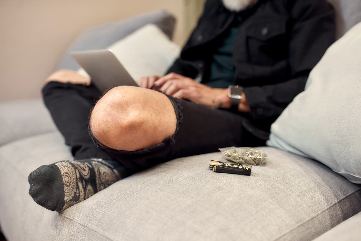 Close up of buds in plastic bag and lighter lying on the couch. Man using laptop in the background. Cannabis and weed legalization concept. Selective focus. Horizontal shot