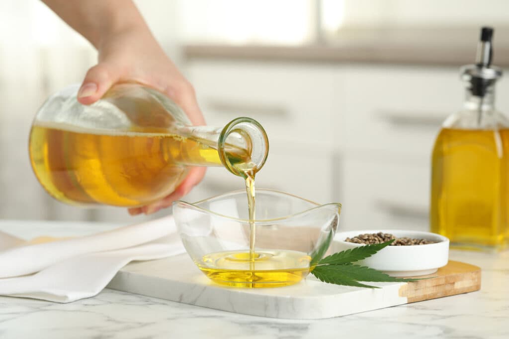 Woman pouring hemp oil into glass bowl at white marble table, closeup