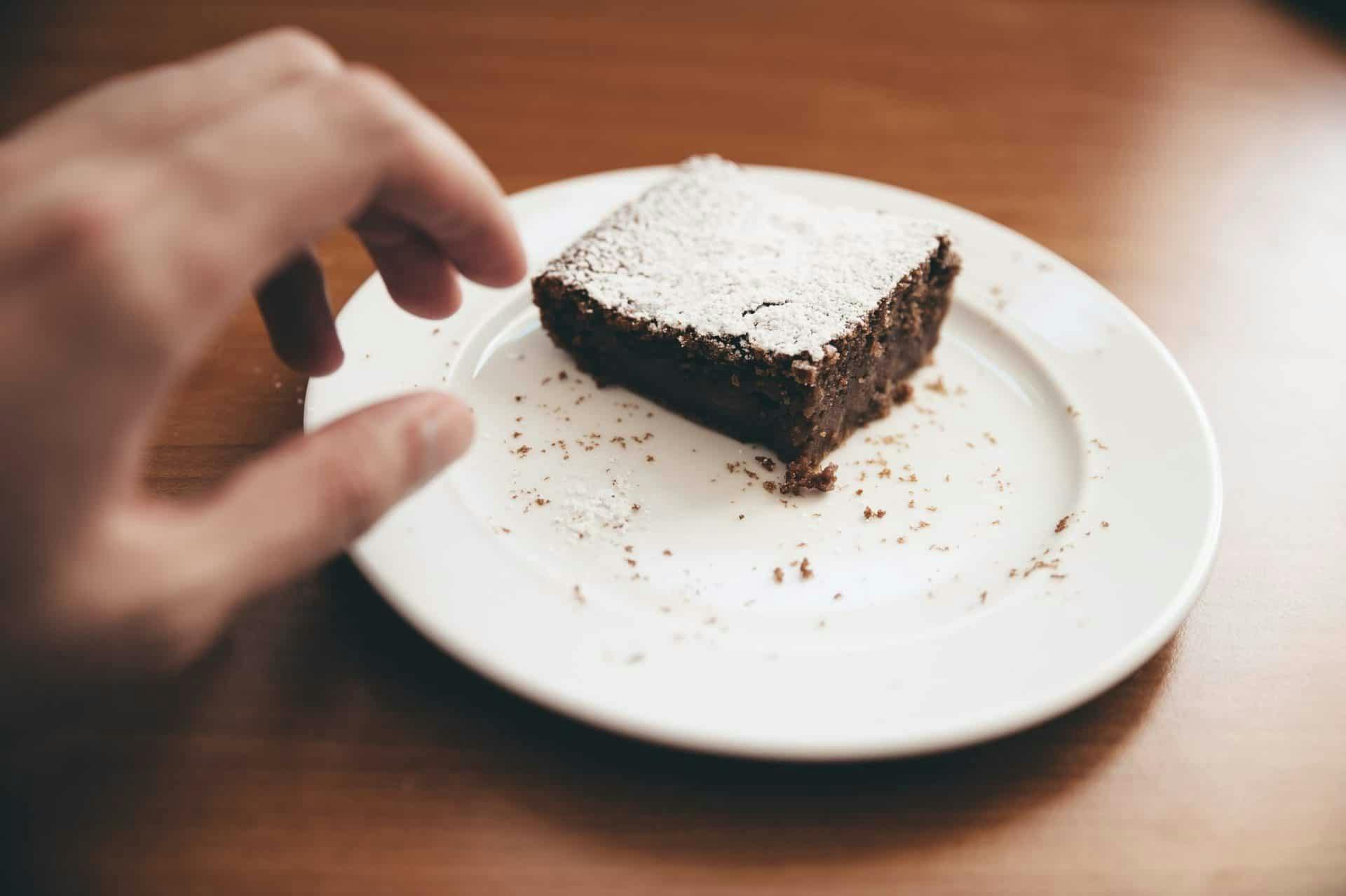 A brownie dusted with powdered sugar sits on a plate, while a hand reaches toward it with delight? Or is it trepidation? By Glenn Carstens via Unsplash