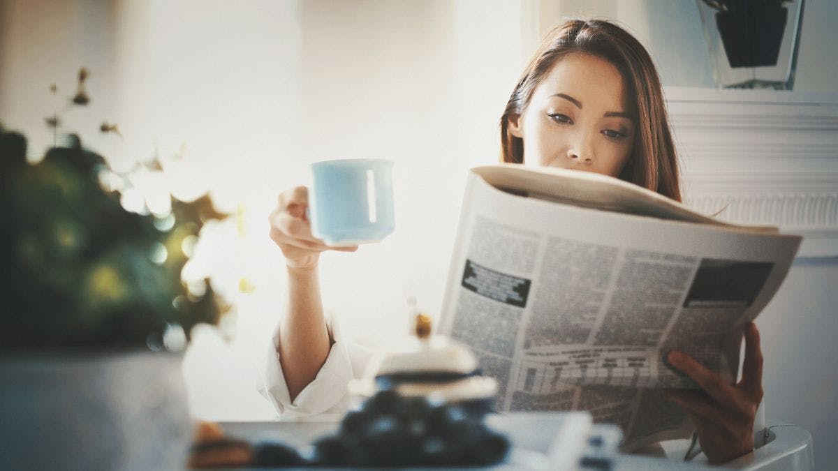 A woman holds a cup of coffee as she reads the newspaper, by Gilaxia via iStock