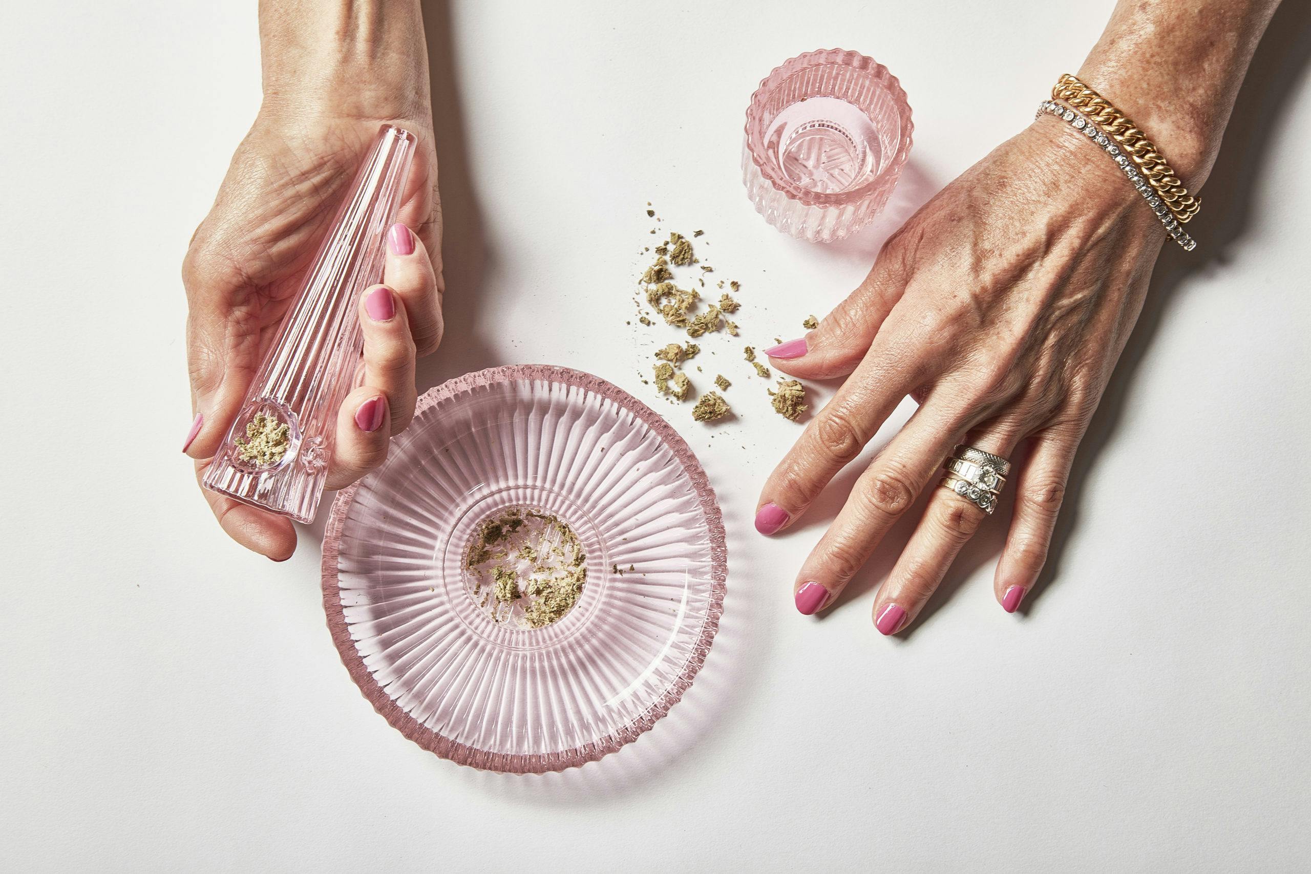 Feminine hands are holding a clear pink pipe with cannabis next to a matching pink ash tray and jar