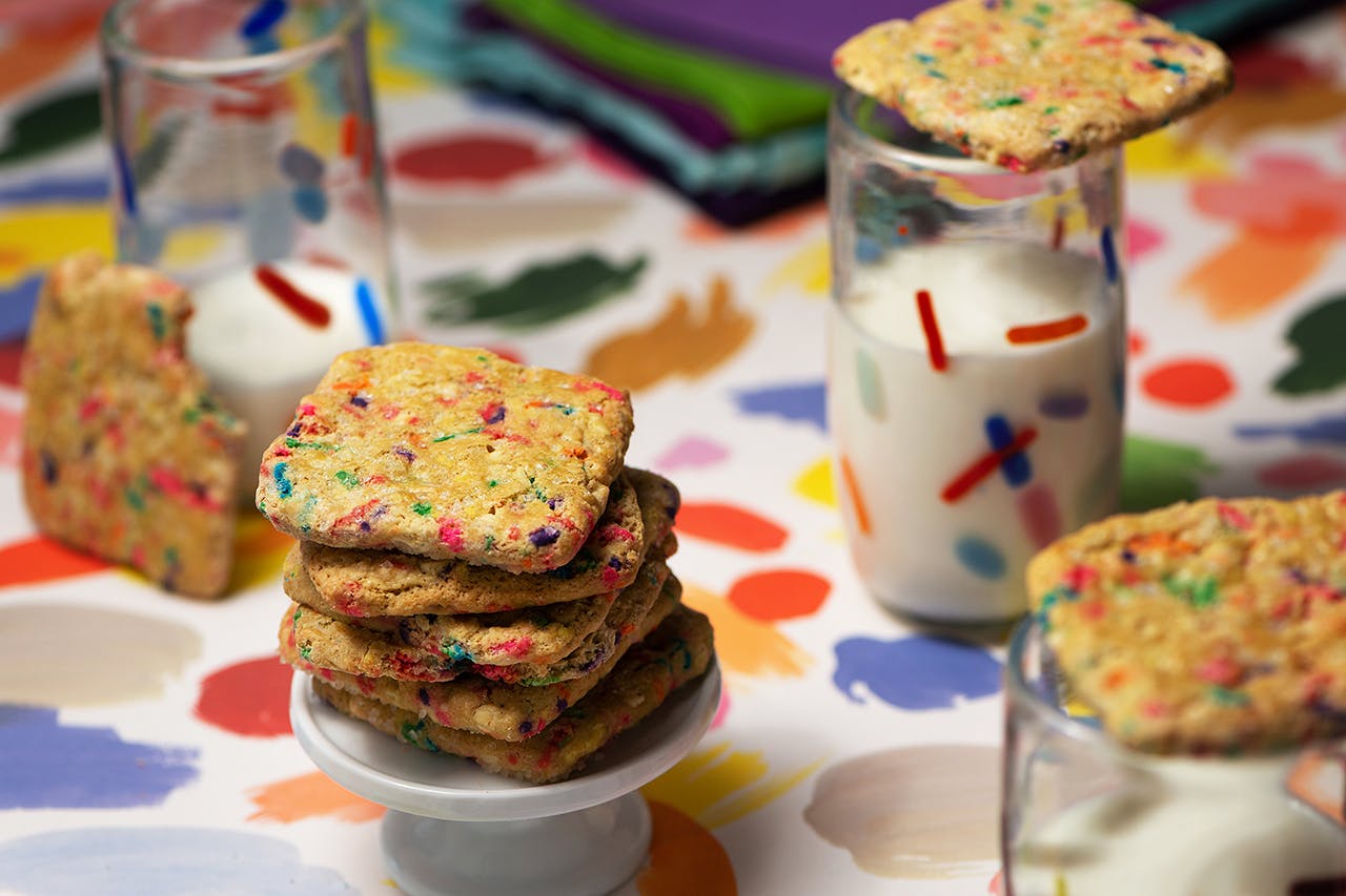 A stack of infused sprinkle sugar cookies on a colorful tablecloth and glasses of milk.