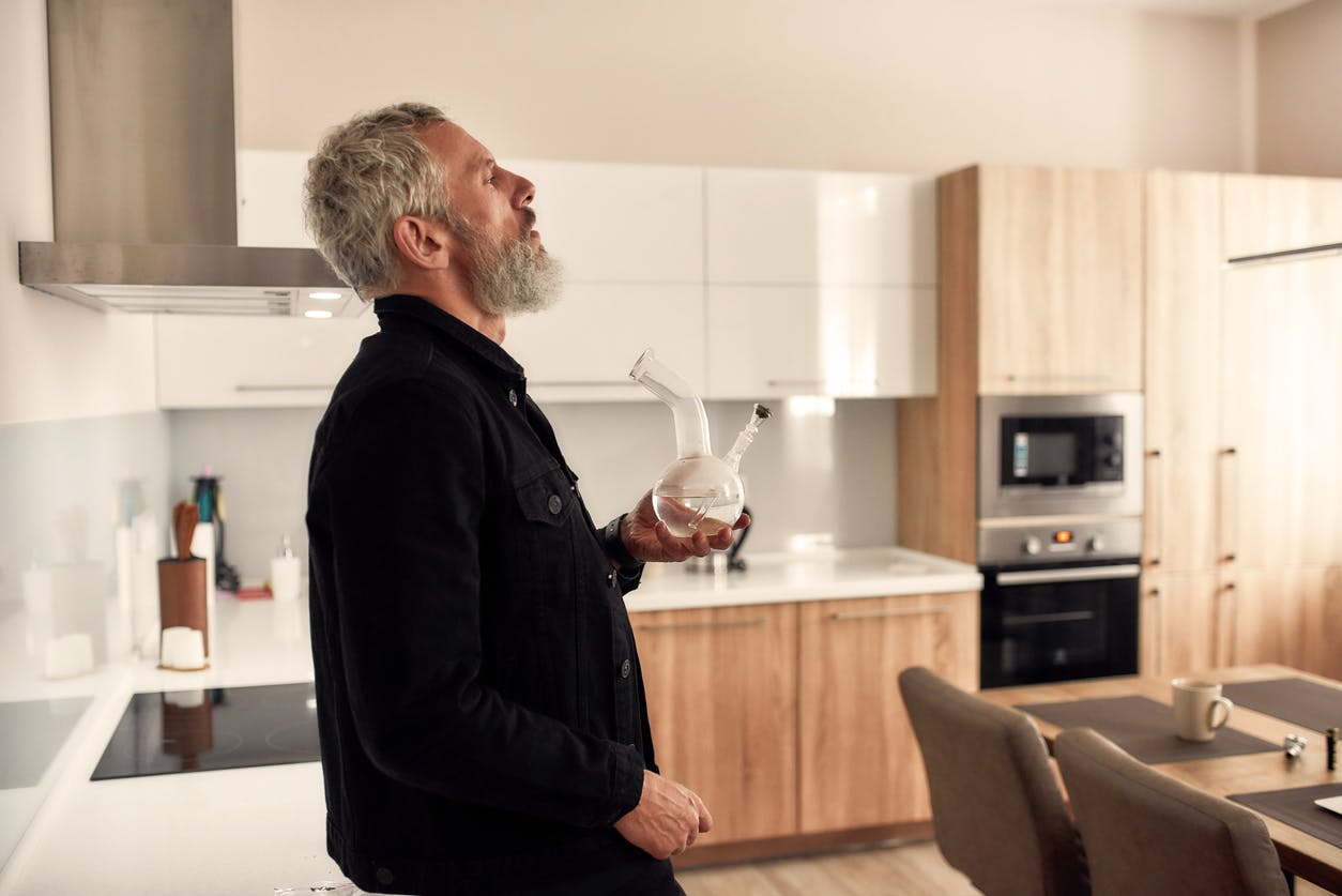 Bearded midle-aged man holding a bong or glass water pipe while smoking marijuana, standing in the kitchen. Cannabis and weed legalization concept. Side view. Horizontal shot