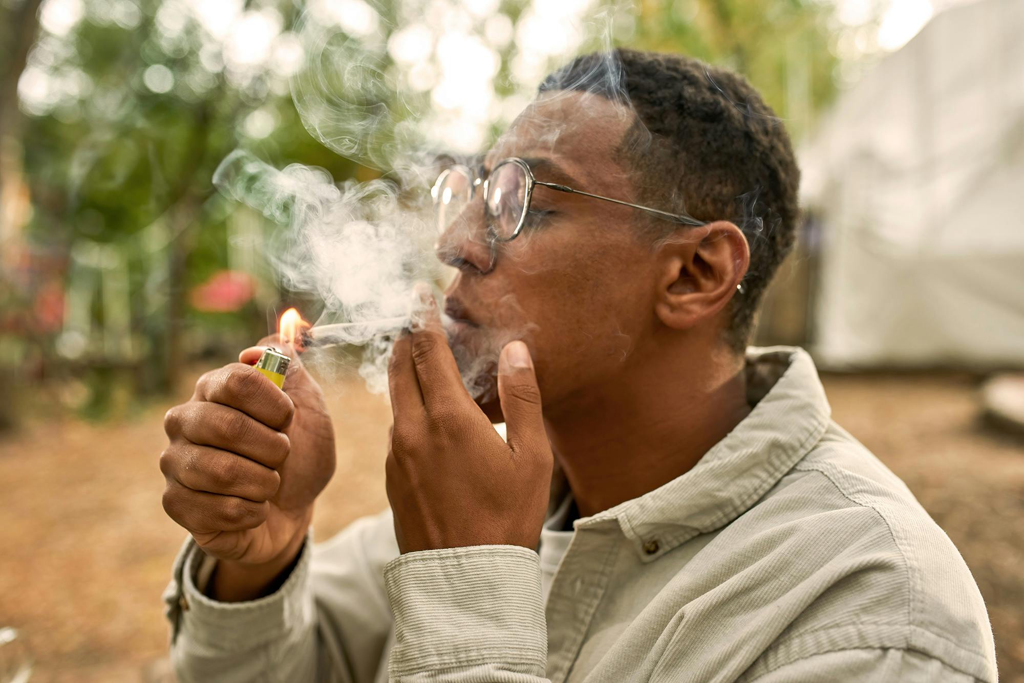Close view of young guy lighting a joint, wearing grey shirt and glasses, smoking on the fresh air , a lot of smoke on the picture, holding a yellow lighter in hand