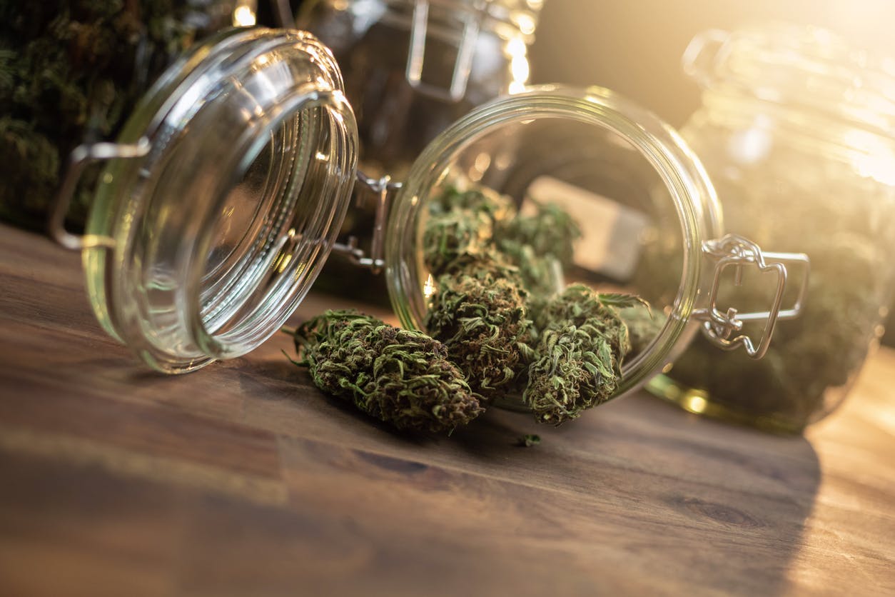 Glass jar with cannabis weed buds spilled on a table with smoke and lens flare.