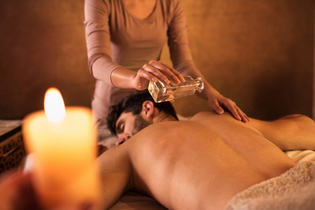 Unrecognizable female massage therapist pouring oil on man's back before massage. Focus is on her hand holding bottle with massage oil.