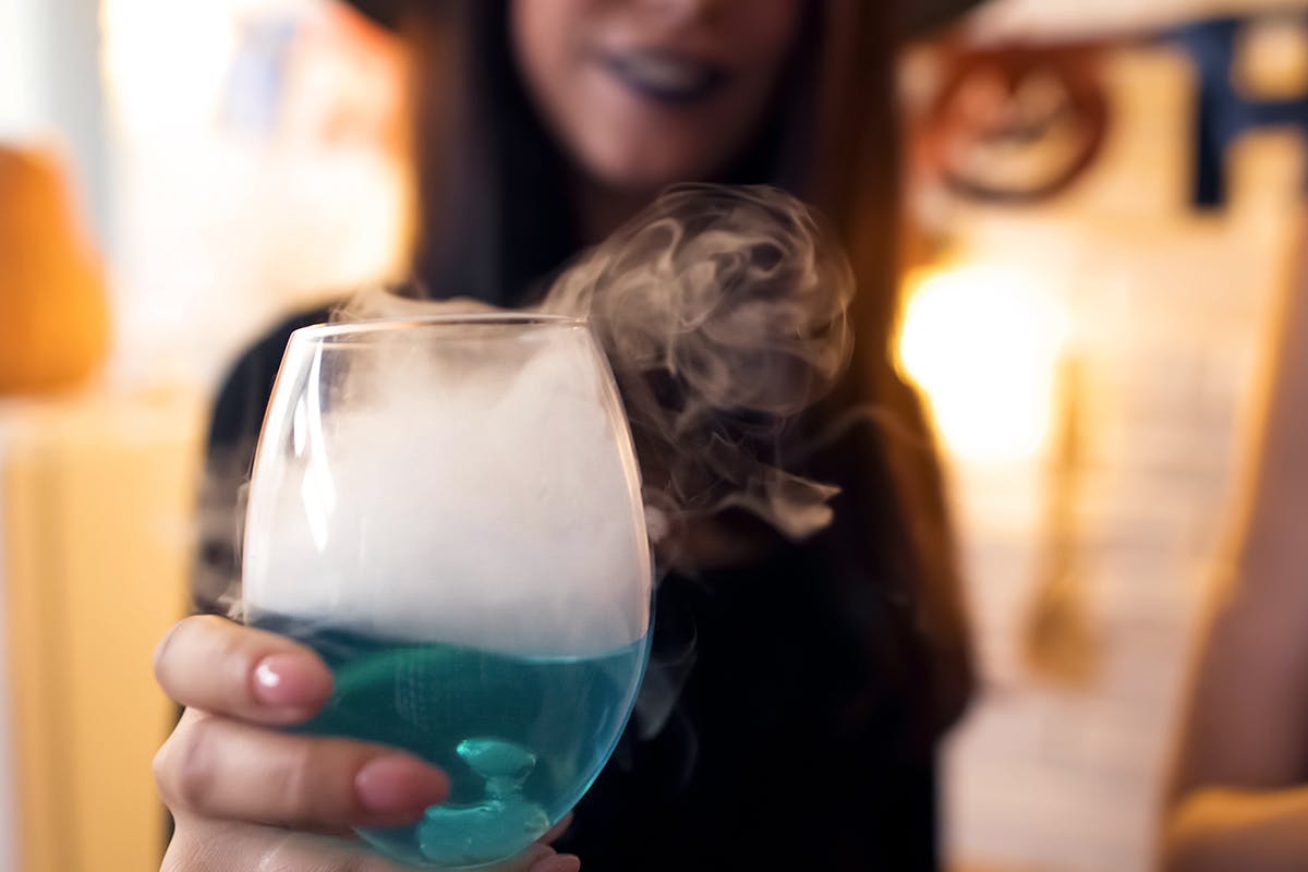 Witch showing blue cocktail with white smoke at Halloween party, having fun