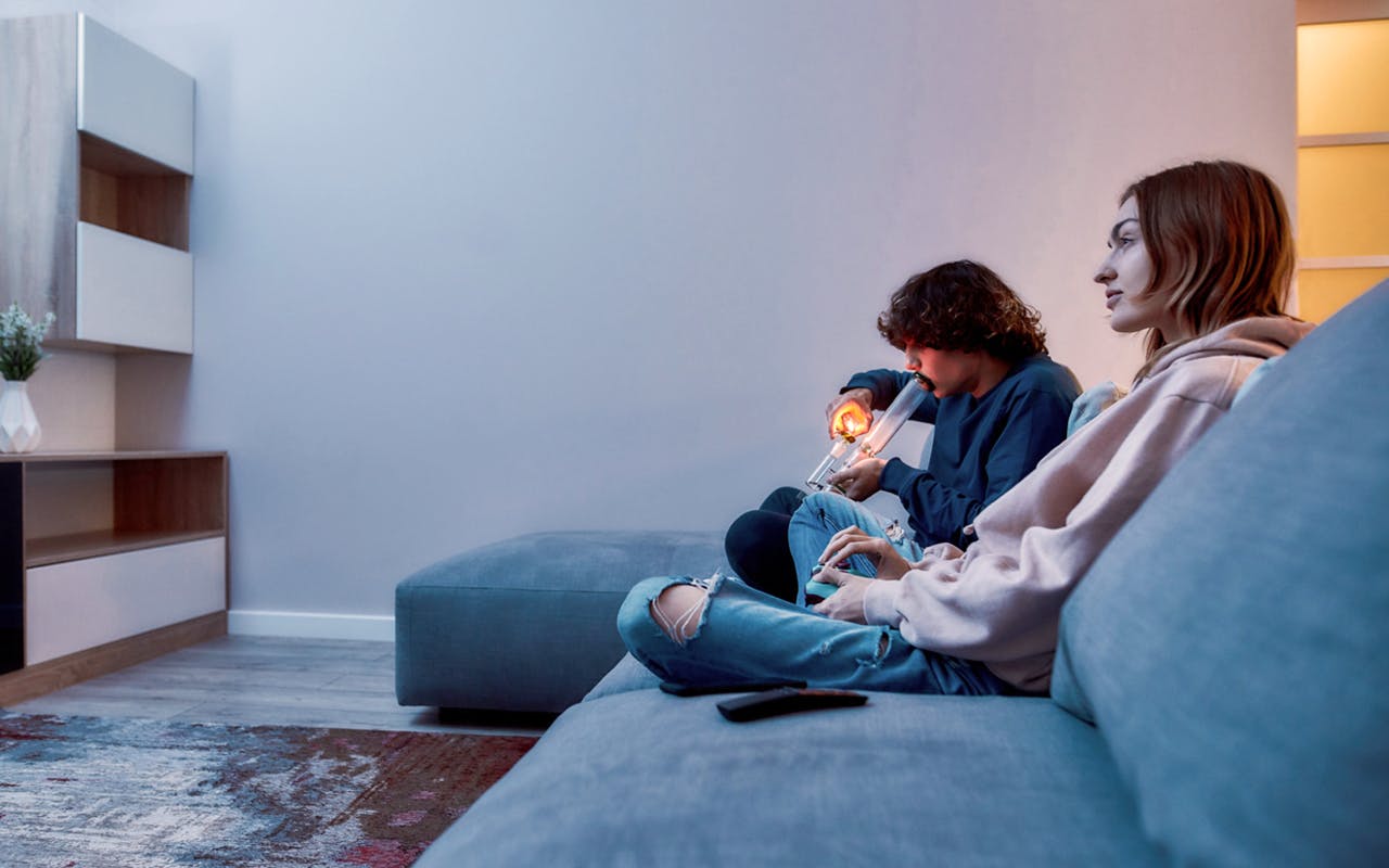 Side view of young caucasian guy lighting cannabis in the bowl of glass water pipe or bong while playing video games with friend, they sitting on the couch at home