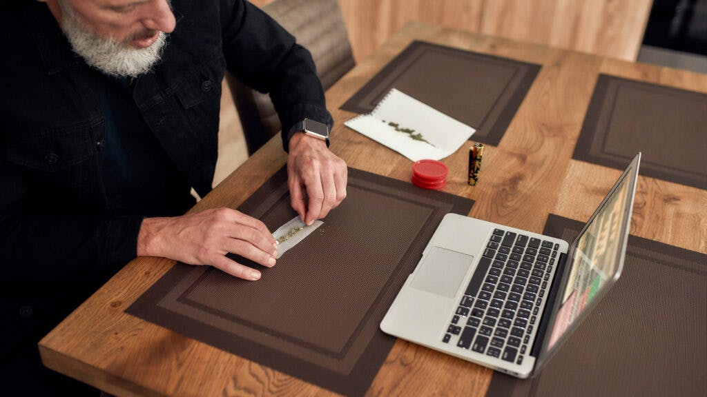 Middle-aged man, artist making marijuana cigarette or joint, sitting at home and writing song using laptop. Marijuana grinder, lighter and weed on the table. Cannabis legalization concept. High angle