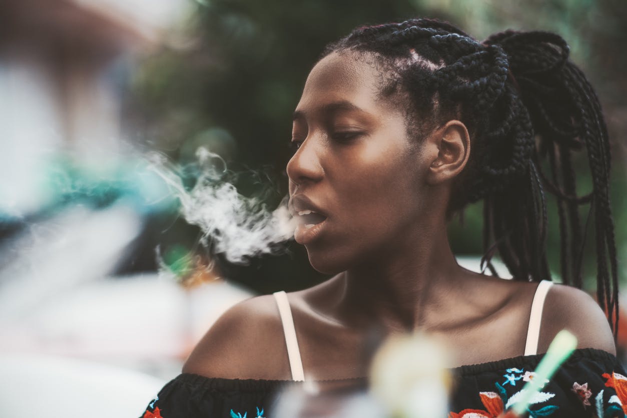 Portrait of a dazzling African girl with braids sitting in a street cafe and exhaling smoke from the hookah; young black female outdoors in a bar plays with the vapor