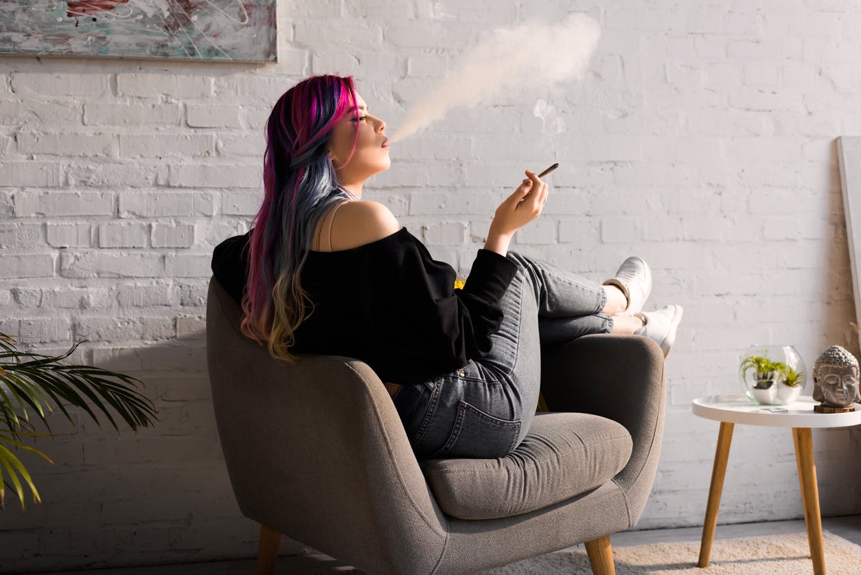 side view of hipster girl with colorful hair sitting in armchair and blowing smoke