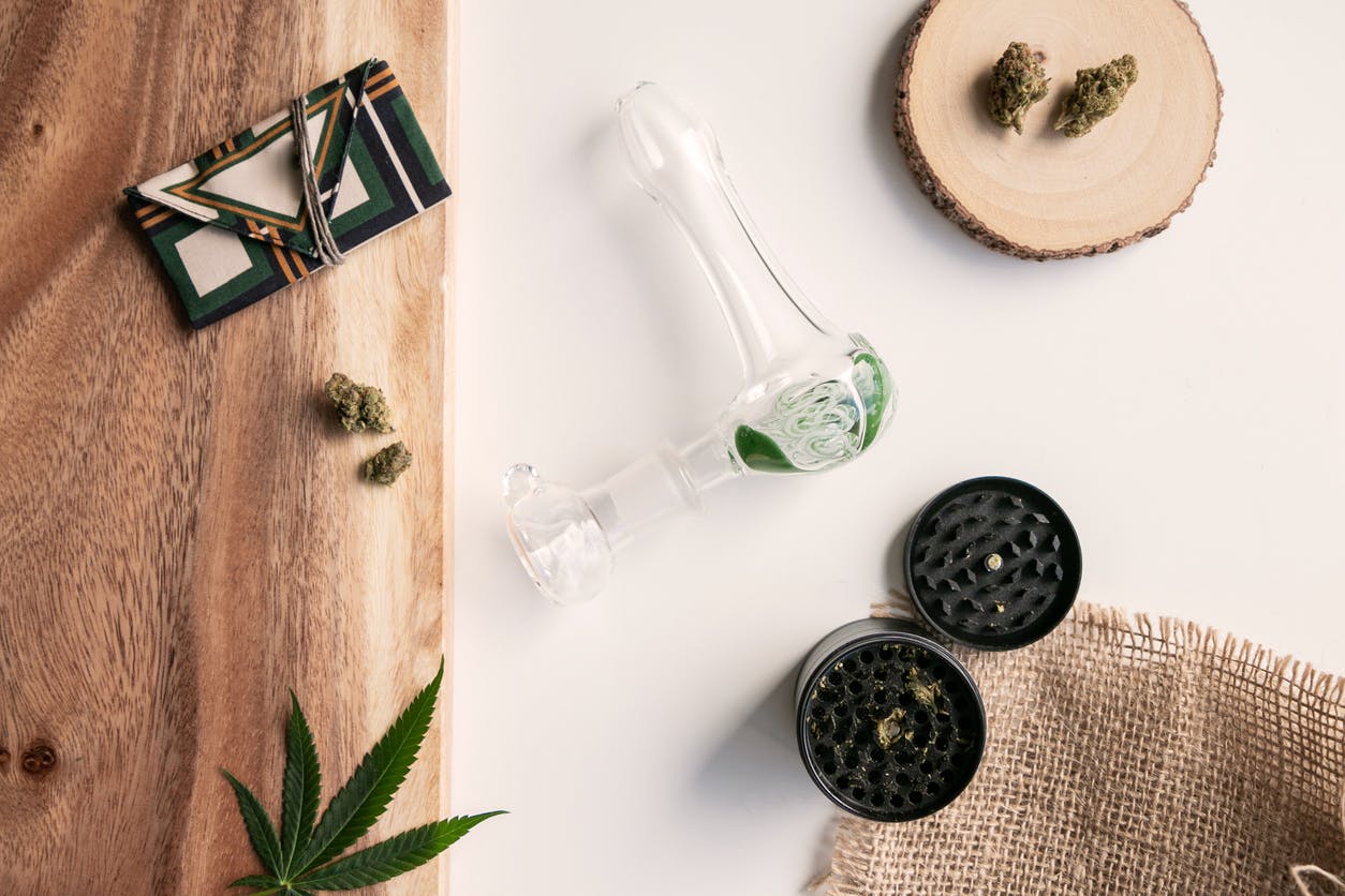 Cannabis herb and wood setting with plaid green weed pouch, nugs, grinder, glass pipe and marijuana leaf on top of wood and white background.